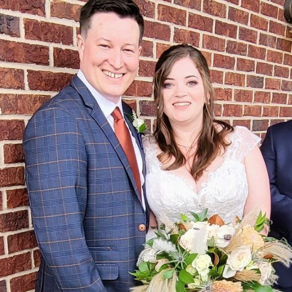 Amanda and Evan smiling on their wedding day, bride holding a bouquet of cream and dried-flower blooms against a brick wall